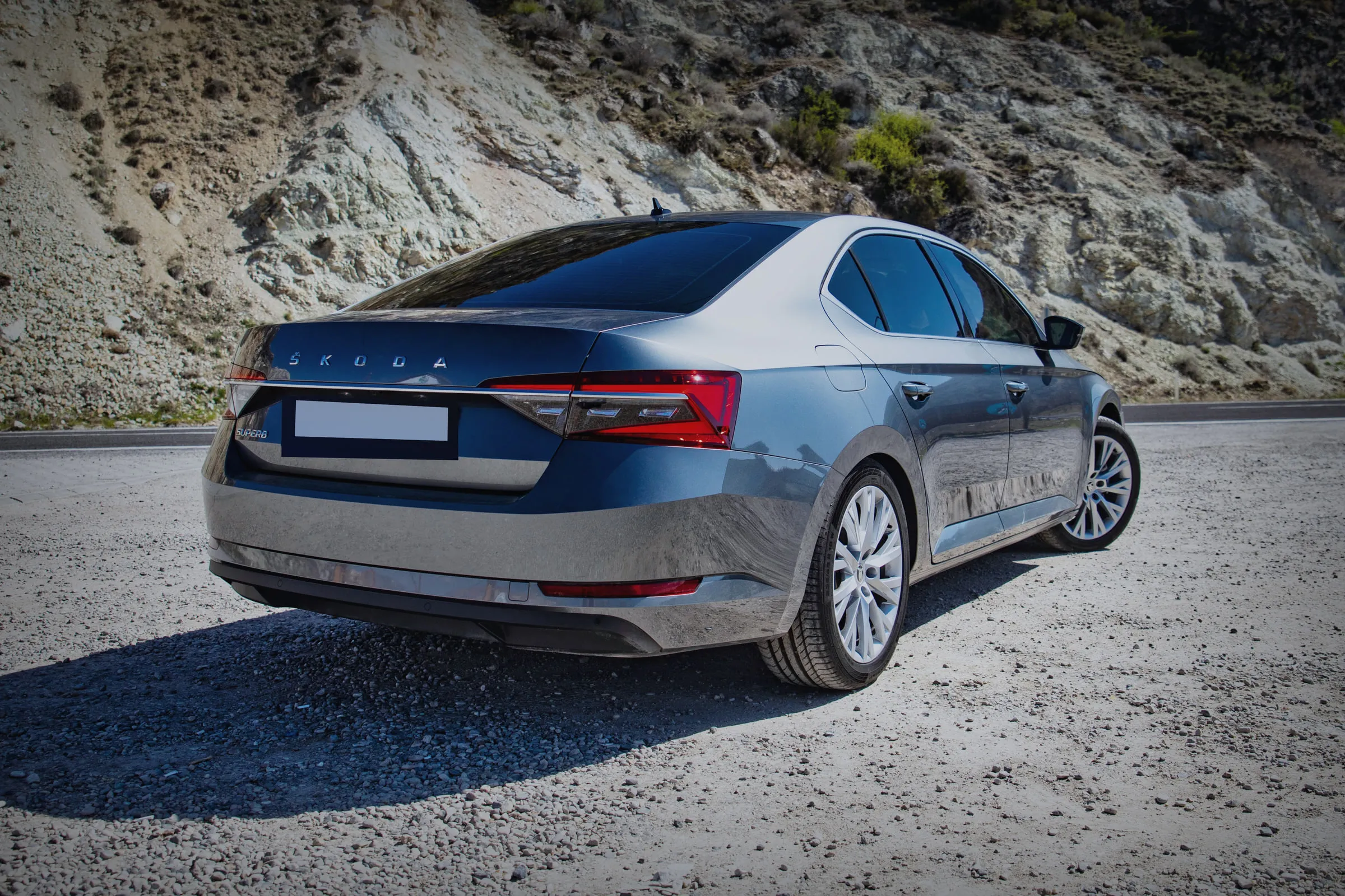 Истата сива Škoda Superb гледана одзади и од страна, на планински пат.